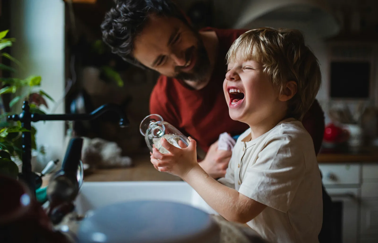 Father laughing with his son at the kitchen sink while filling a glass of water — the water families drink every day can carry documented endocrine disruptors like PFAS, lead, and arsenic