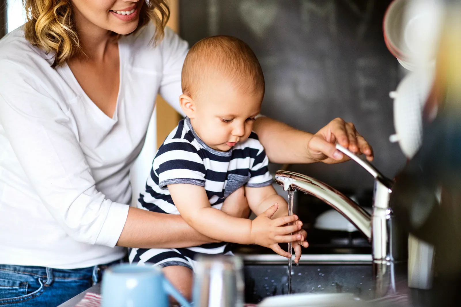 Mother helping baby reach toward a kitchen faucet with running water